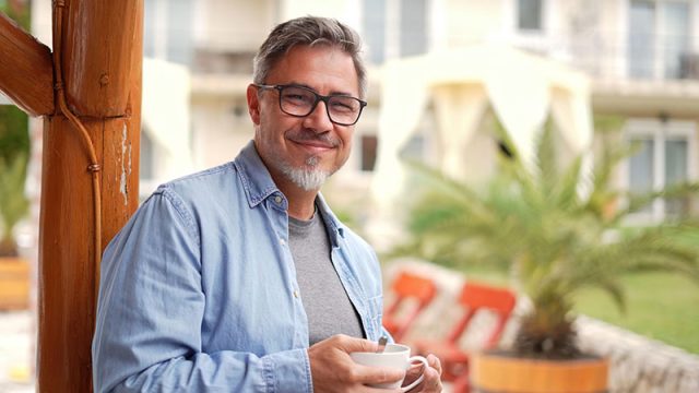 Man smiling while holding a coffee cup, showcasing a confident smile, in a relaxed outdoor setting with palm trees and modern architecture, relevant to discussions on smile problems and porcelain veneers at Algoma Family Dentistry.
