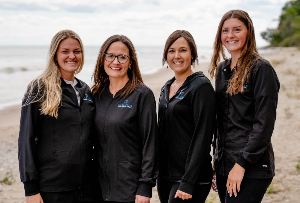Four dental assistants from Algoma Family Dentistry smiling together on a beach, wearing black uniforms with the practice logo, showcasing a friendly and approachable team atmosphere.