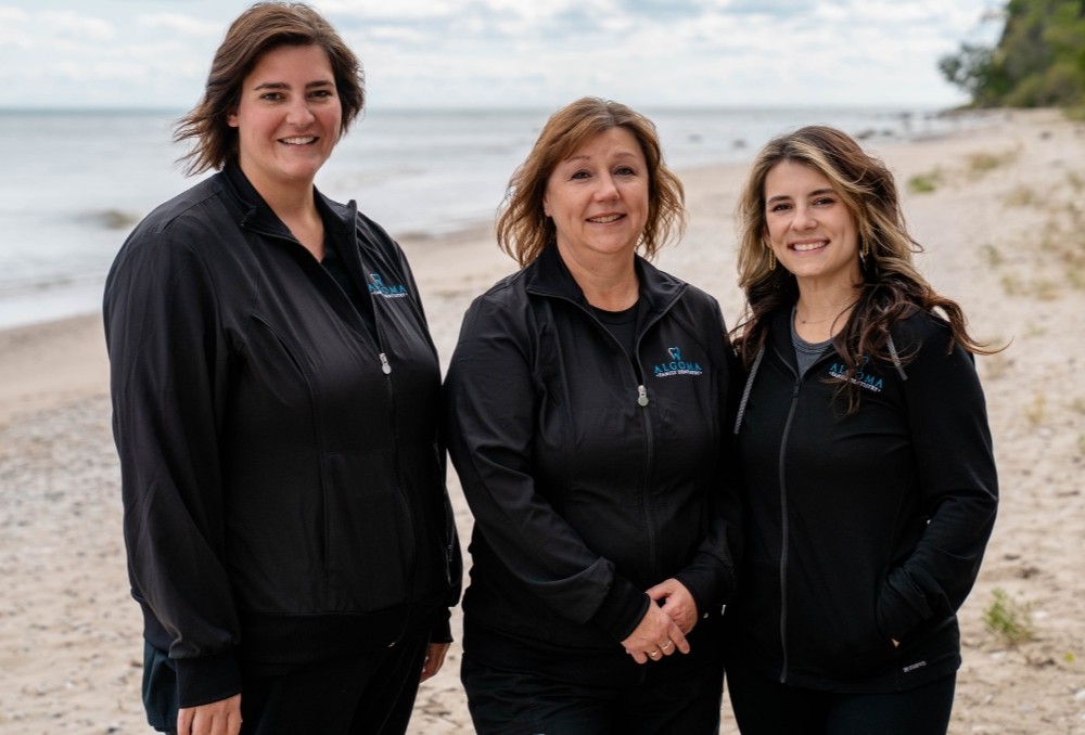 Lisa, Kayla, and Amber from Algoma Family Dentistry, standing on a beach, wearing black jackets with the practice logo, smiling and showcasing their friendly demeanor.