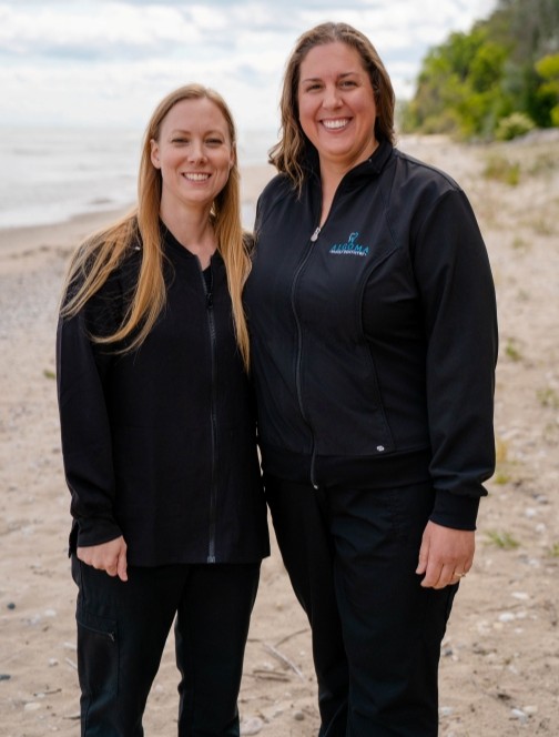 Emily and Barb, dental hygienists at Algoma Family Dentistry, standing together on a beach, smiling, wearing black dental attire.